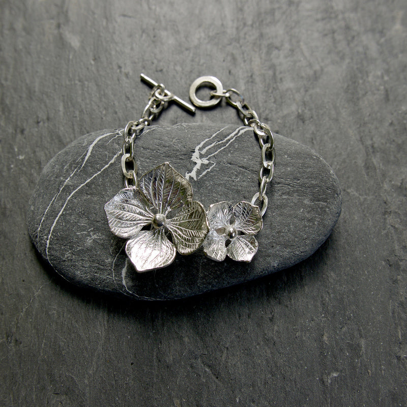 A handmade Sterling silver hydrangea flower bracelet. This very pretty floral bracelet has two flowers sitting across your wrist with an oval chain and a T-bar catch. Hallmarked botanical jewellery, made in the UK.